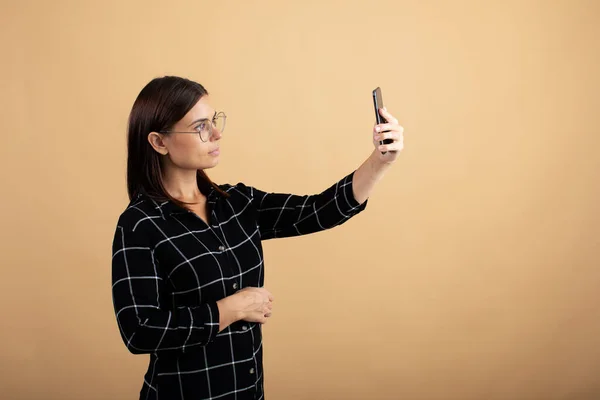 Молодая женщина в клетчатом платье стоит на оранжевом фоне и играет со своим телефоном — стоковое фото