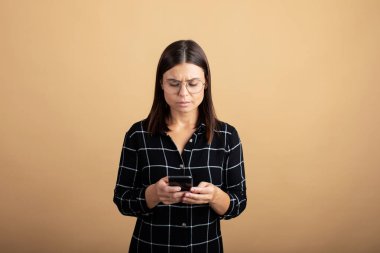 Ekose elbiseli genç bir kadın turuncu arka planda duruyor ve telefonuyla oynuyor.