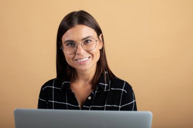 Ekose elbiseli genç bir kadın turuncu bir arka planda dizüstü bilgisayarla duruyor.