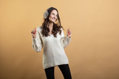 İzole edilmiş turuncu bir arka planda beyaz kazaklı, poz veren ve gri kulaklıklı güzel bir kadının görüntüsü.