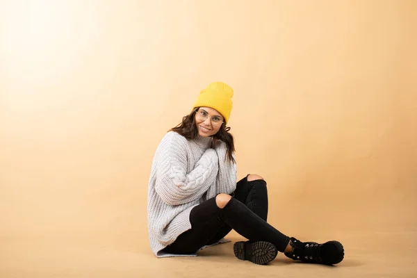 Una joven vestida con un suéter, sombrero amarillo y gafas se sienta en el suelo sobre un fondo naranja y sueña — Foto de Stock