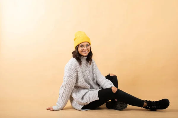 Молодая женщина в свитере, желтой шляпе и очках сидит на полу на оранжевом фоне и мечтает — стоковое фото