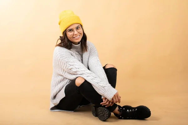 Молода жінка, одягнена в светр, жовтий капелюх і окуляри сидить на підлозі на помаранчевому фоні і мріє — стокове фото