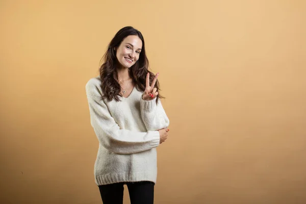 Изображение молодой красивой женщины в белом свитере, позирующей изолированно на фоне оранжевой стены и показывающей 2 пальца — стоковое фото