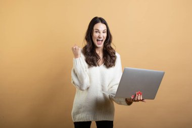 İzole edilmiş turuncu bir arkaplanda dizüstü bilgisayarla poz veren beyaz kazaklı güzel bir kadının görüntüsü.