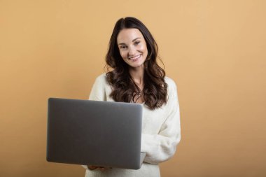 İzole edilmiş turuncu bir arkaplanda dizüstü bilgisayarla poz veren beyaz kazaklı güzel bir kadının görüntüsü.