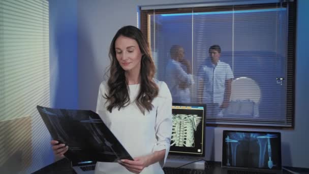 Uma foto de uma bela jovem médica. O médico está de pé na sala de tomografia e estudando a imagem do raio-x, ela sorri encantadoramente e olha para a câmera. Profissionais no trabalho . — Vídeo de Stock