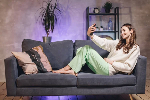 Una joven sentada en un sofá en una acogedora habitación se toma una selfie y sonríe — Foto de Stock