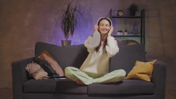 Mladá žena sedící na pohovce v útulné místnosti a poslouchající hudbu na bezdrátových sluchátkách. Dívka šťastná. — Stock video