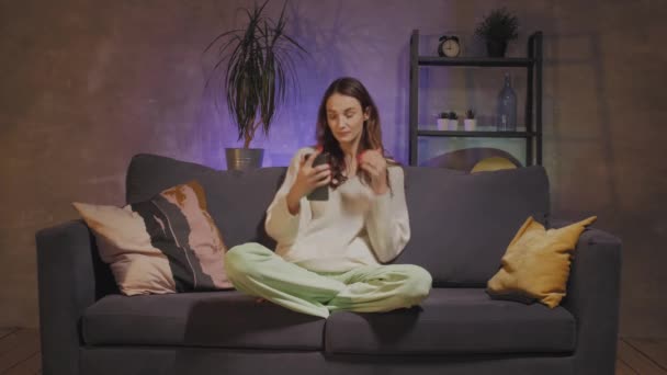 Mladá žena sedící na pohovce v útulném pokoji si vezme selfie a usměje se — Stock video