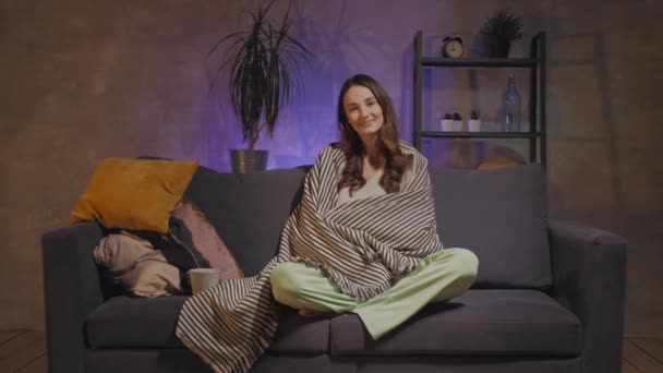 A young woman sits on a sofa in a cozy room wrapped in a plaid, drinking tea and watching TV with her son — Stock Video