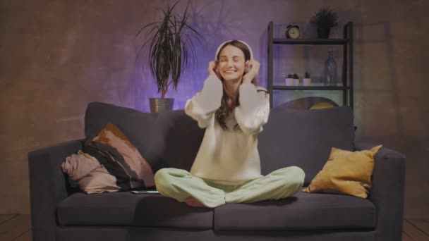 Een jonge vrouw zittend op een bank in een gezellige kamer luisterend naar muziek op een draadloze hoofdtelefoon. Meisje gelukkig. — Stockvideo