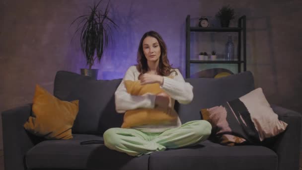 Молодая женщина сидит на диване в уютной комнате и смотрит телевизор — стоковое видео