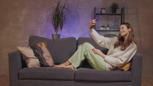 En ung kvinna som sitter i en soffa i ett mysigt rum tar en selfie och ler — Stockvideo