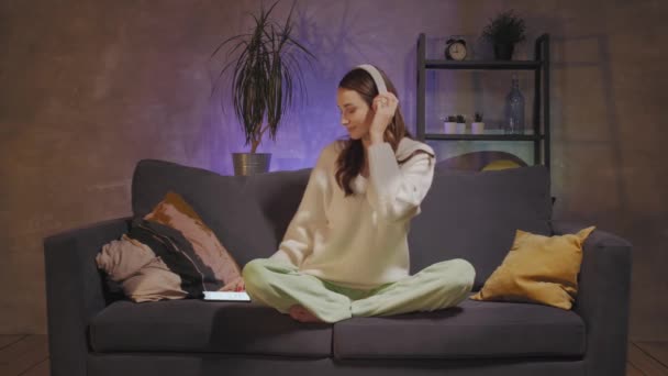 Una joven sentada en un sofá en una acogedora habitación escuchando música con auriculares inalámbricos. Chica feliz . — Vídeo de stock