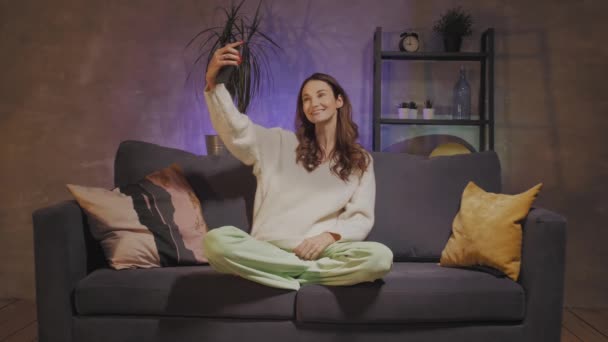 Eine junge Frau, die in einem gemütlichen Zimmer auf einem Sofa sitzt, macht ein Selfie und lächelt — Stockvideo