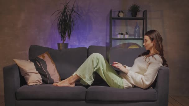 En ung kvinna som sitter i en soffa i ett mysigt rum och arbetar med en bärbar dator — Stockvideo