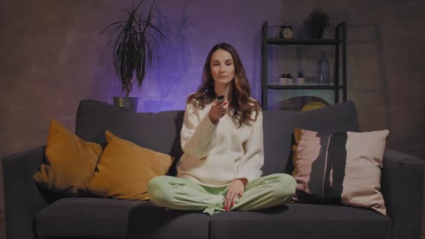 Una joven está sentada en un sofá en una acogedora habitación viendo la televisión — Vídeos de Stock