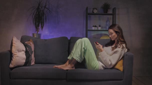 Ung mor och son sitter på soffan i ett mysigt rum och leker på telefonen — Stockvideo