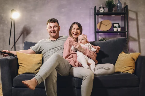 Familia joven sentada en el sofá en una habitación acogedora. Mamá, papá y bebé son felices. — Foto de Stock