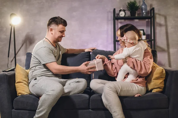 Familia joven en un acogedor apartamento en el sofá. La familia es feliz. El marido le da un regalo a su esposa. . — Foto de Stock