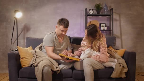 A young family sits on a sofa in a cozy room. Mom, dad and baby reading a book. Family happy — Stock Video