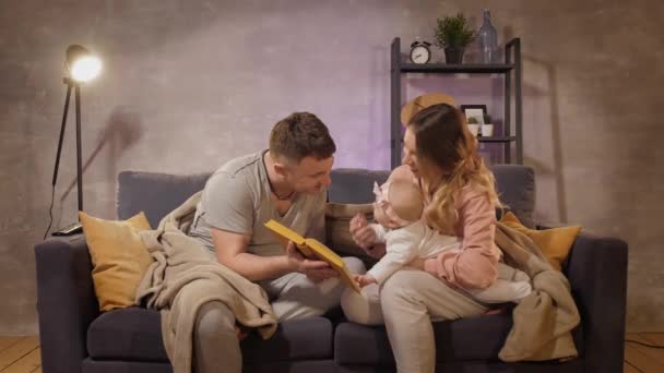 En ung familie sitter på en sofa i et koselig rom. Mamma, pappa og baby leser en bok. Familieglad – stockvideo