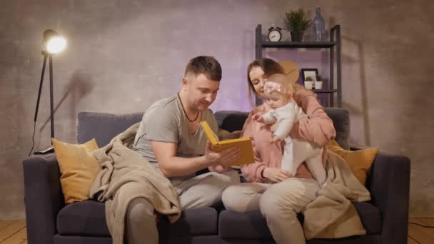 Eine junge Familie sitzt auf einem Sofa in einem gemütlichen Zimmer. Mama, Papa und Baby lesen ein Buch. Familie glücklich — Stockvideo