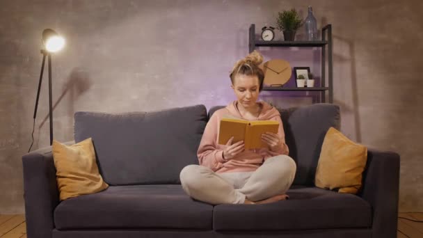 Mladá žena sedí na pohovce a čte si knihu.. — Stock video