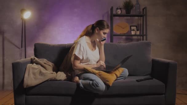 Giovane bella ragazza seduta sul divano a lavorare su un computer portatile. Ragazza che parla al telefono — Video Stock