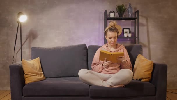 Una joven está sentada en el sofá leyendo un libro. El hombre hizo un regalo a su amado . — Vídeo de stock