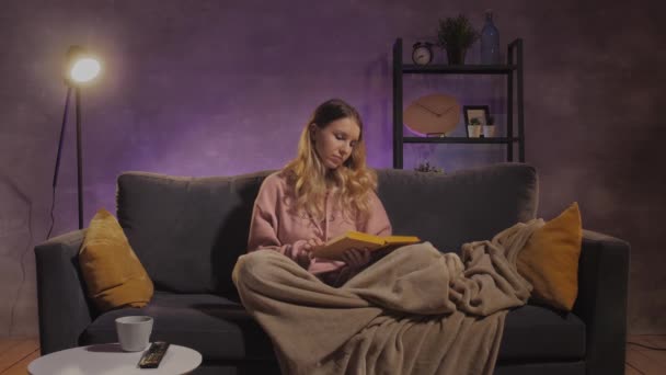 A young woman is sitting on the couch reading a book. Girl dreams on a cozy couch in a warm room — Stock Video