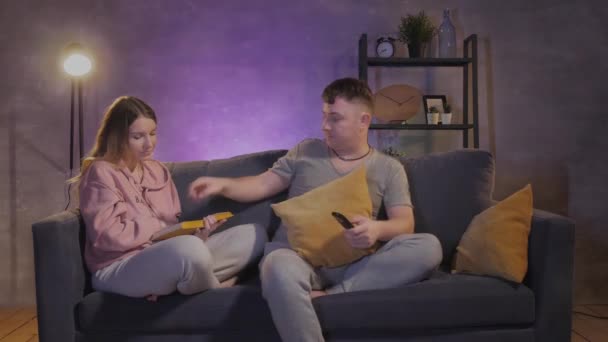 Un hombre ve televisión. Hermosa chica leyendo un libro cerca de su marido — Vídeo de stock