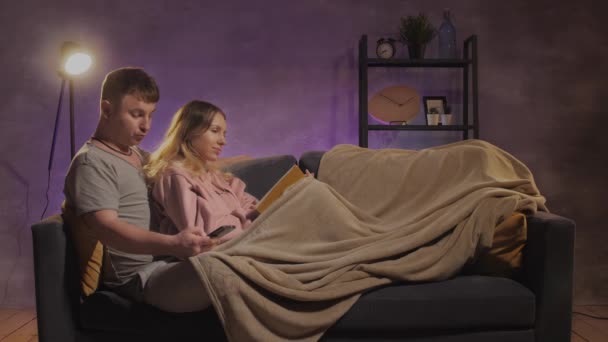 En ung kvinna och en man sitter i en soffa och läser en bok. En kille och en flicka lycklig — Stockvideo