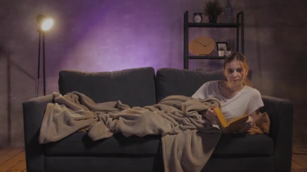 Joven chica hermosa acostada en el sofá leyendo un libro. Chica viendo la televisión — Vídeos de Stock