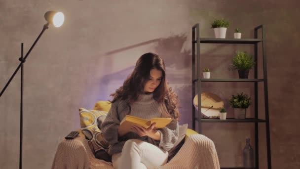 若い女性が本を読んで椅子に座っている。女の子は暖かい部屋で居心地の良いソファで夢を見る — ストック動画
