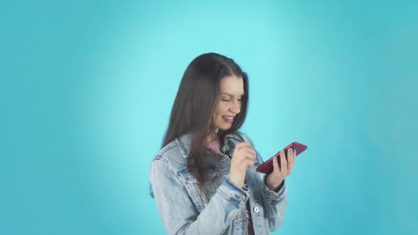 Mujer joven en chaqueta de mezclilla escribe un mensaje en el teléfono inteligente sobre fondo azul — Vídeo de stock