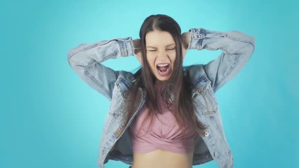 Молодая женщина в джинсовой куртке с руками за ушами кричит на голубом фоне — стоковое видео