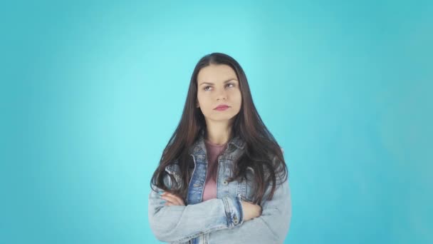 Una joven descontenta con una chaqueta de mezclilla sobre un fondo azul mira a la cámara — Vídeos de Stock