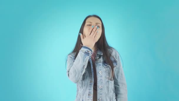 Joven mujer cansada en una chaqueta de mezclilla bosteza en la cámara sobre un fondo azul — Vídeo de stock