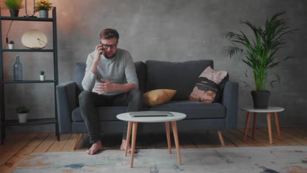 Jeune homme à lunettes en colère parlant au téléphone assis sur le canapé. Jeune homme d'affaires . — Video