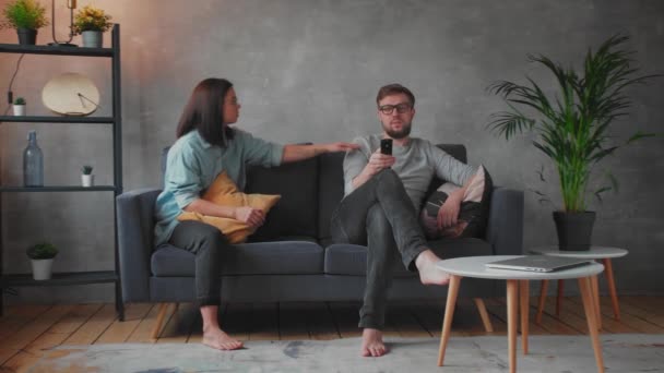 Jeune couple assis à la maison sur le canapé. Homme regarde la télé. Jeune femme appelle son mari, il n'entend pas. Un petit enfant joue devant la télé . — Video