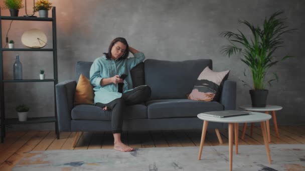 Uma jovem está sentada no sofá e a enviar mensagens. A mulher joga o telefone no sofá e cobre seu rosto. Vídeo social — Vídeo de Stock