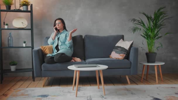 Mujer joven con gafas enojada hablando por teléfono sentada en el sofá. Joven empresaria . — Vídeo de stock