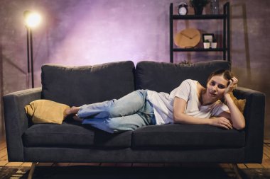 Kanepede oturan bir kadın rahat bir odada TV seyrediyor ve ekranda gördüklerine üzülüyor.
