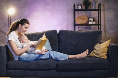 Genç bir kız bebeğiyle kanepede uzanıp kitap okuyor. Samimi oda