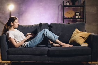 Young girl lying on the couch and working on a laptop. Cozy room