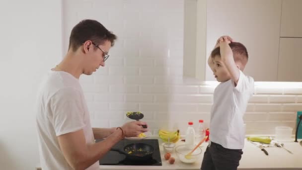 Niño hijo ayuda a un joven padre feliz preparar la comida en una cocina brillante — Vídeo de stock