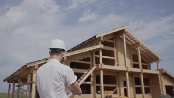 Bauarbeiter mit Helm steht mit dem Rücken zu einem Holzhaus — Stockvideo