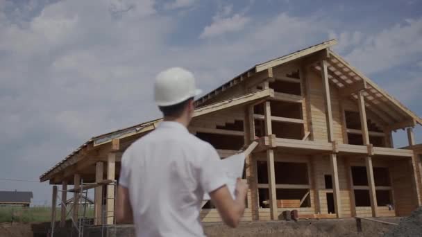 Voorman Bouwer in helm staand met zijn rug naar een houten huis — Stockvideo